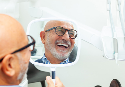 A bald, elderly man smiling while using a hand mirror